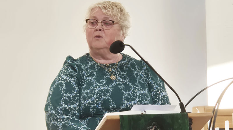 En dame i grønn blomstrete kjole står ved talerstolen og snakker i mikrofonen