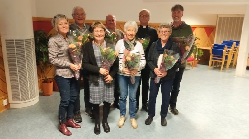 En gruppe på åtte mennesker, fire damer fremme og fire menn bak, de står og smiler og holder hver sin bukett med blomster