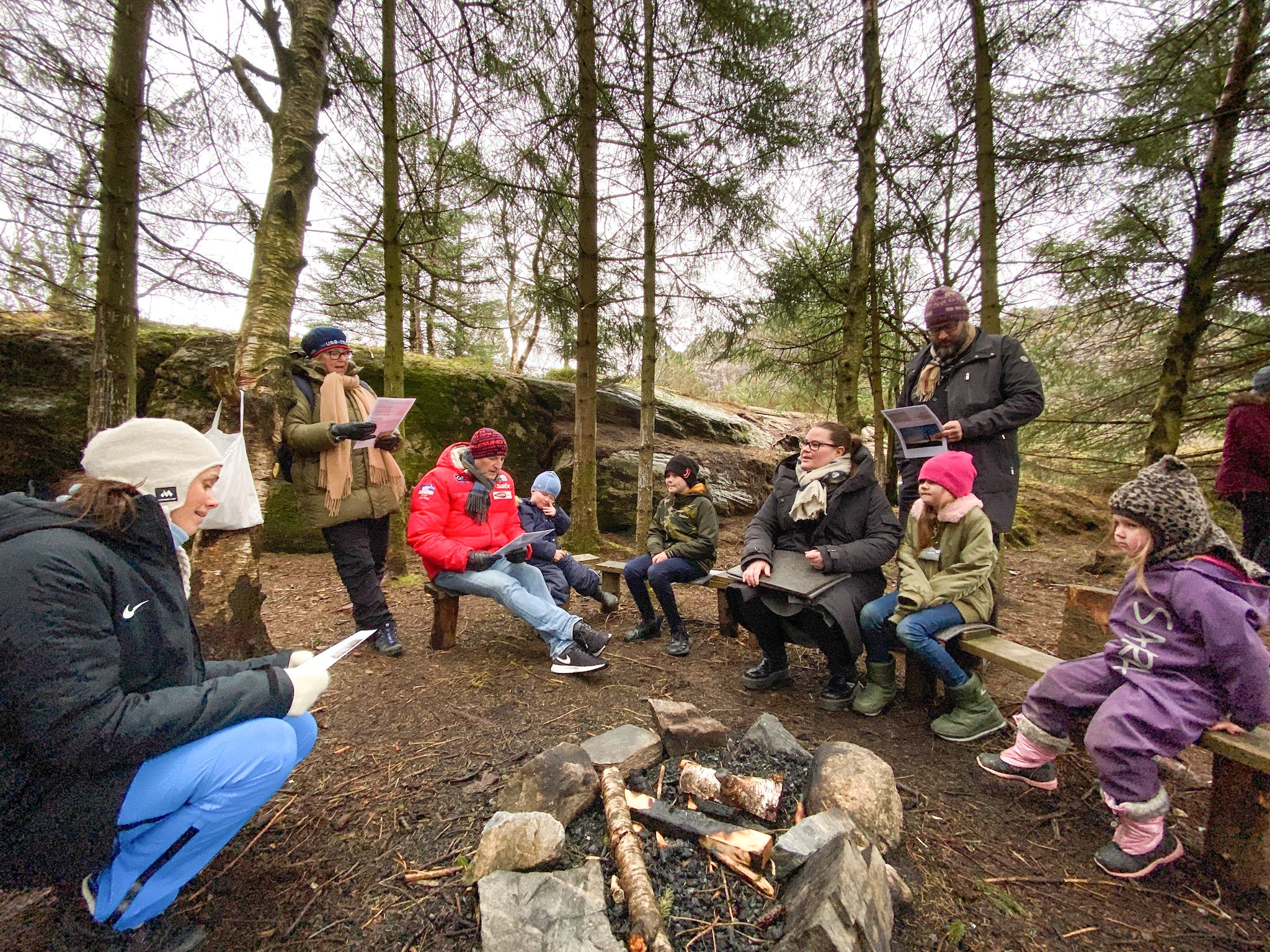 Barn og voksne sitter ute på tømmerbenker rundt et bål, de synger en sang