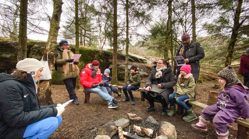 Barn og voksne sitter ute på tømmerbenker rundt et bål, de synger en sang