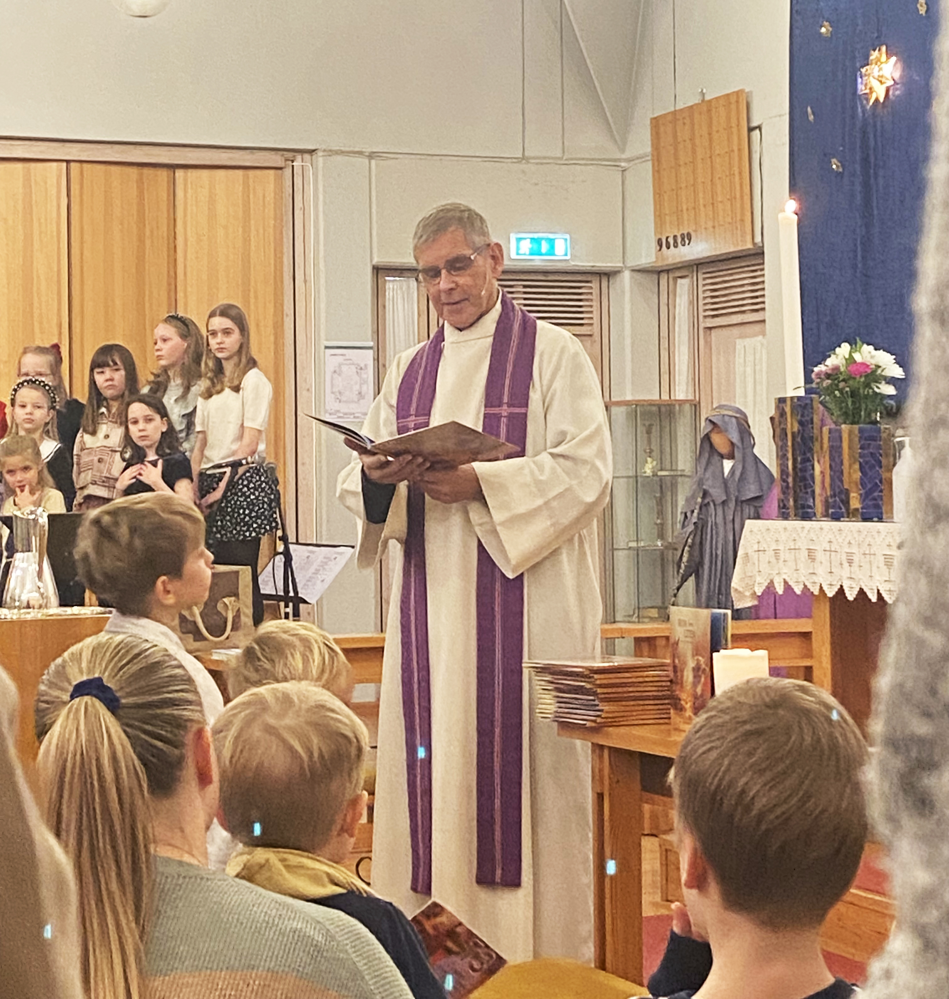 Presten st&aring;r og leser fra juleboka med barn og voksne framfor seg