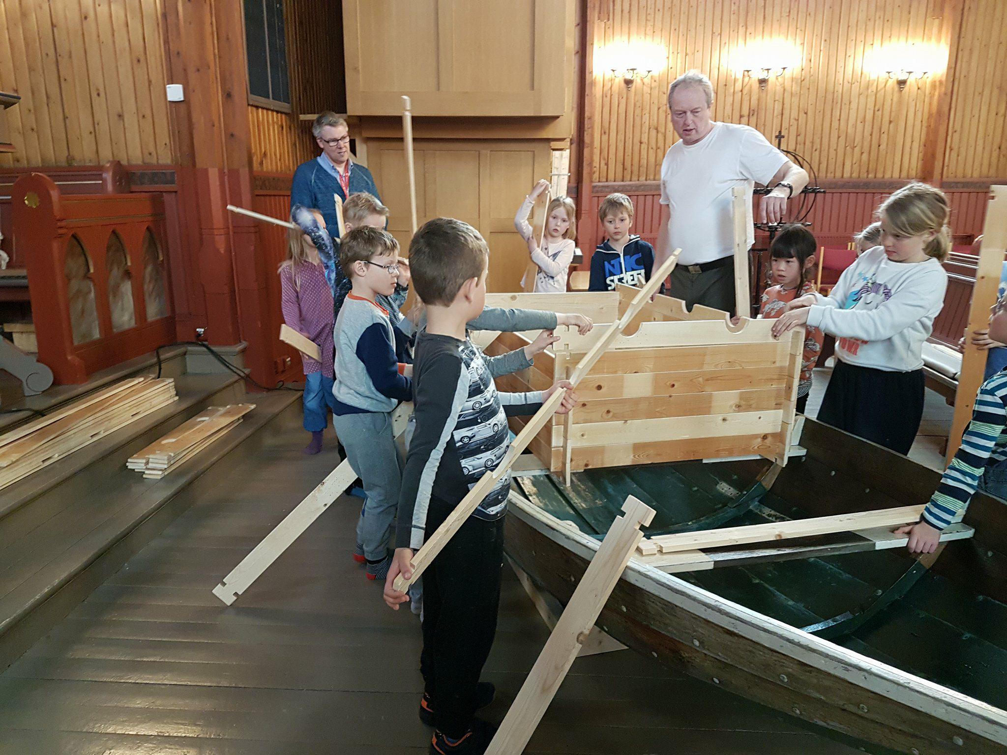 To voksne og mange barn står rundt en robåt (Noahs Ark) og bygger et hus på båten