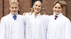 Tre smilende konfirmanter i hvite kapper utenfor en kirke