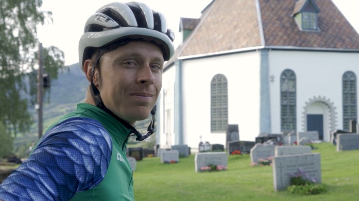 Jonas Orset er syklist og sykler fra kirke til kirke, her utenfor en kirke ikledd sykkelhjelm.
