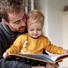 En voksen mann som sitter med et barn på fanget, og leser en bok.